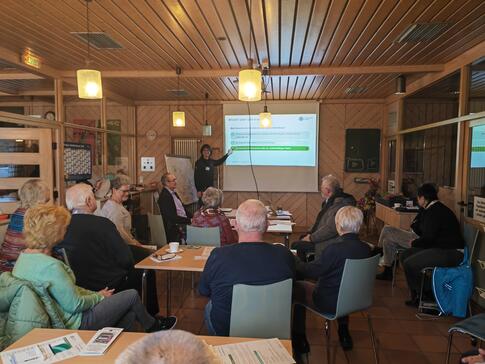 Die Besucher:innen sitzen im Raum und blicken zu Johannes, der mit seinem Finger auf die Leinwand zeigt