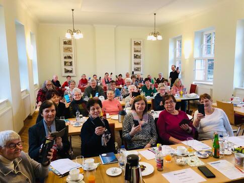 Knapp 50 Senior:innen sitzen an Tischreihen und halten ihr eigenes Smartphone nach oben. Das Bild stammt von der Veranstaltung am Nachmittag.