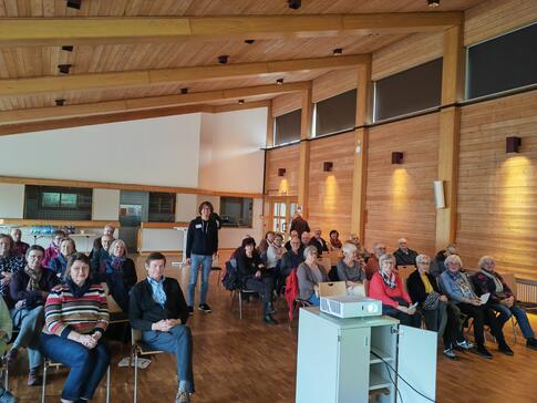 Die Teilnehmenden sitzen auf Stühlen im Veranstaltungsraum und schauen zur Präsentation nach vorne während Johannes in der Mitte des Raumes steht