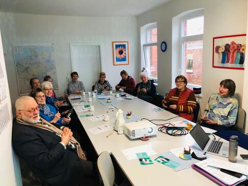 Elf Senior:innen sitzen an einem großen Tisch im Viereck. Einige gucken in die Kamera. Andere halten ein Smartphine in der Hand. Auf dem  Tisch stehen ein Beamer und ein Laptop von Petra.