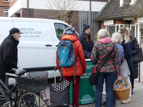 Pera steht in Winterjacke und Mütze vor dem Info-Mobil am Info-Stand. Um den Stand herum stehen fünf Damen mit Einkaufstaschen und ein Herr mit einem Fahrrad.