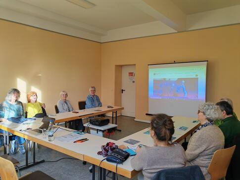 Die Teilnehmenden sitzen auf Stühlen an einem zusammengeschobenen Tisch im Raum und schauen zur Präsentation an der Leinwand