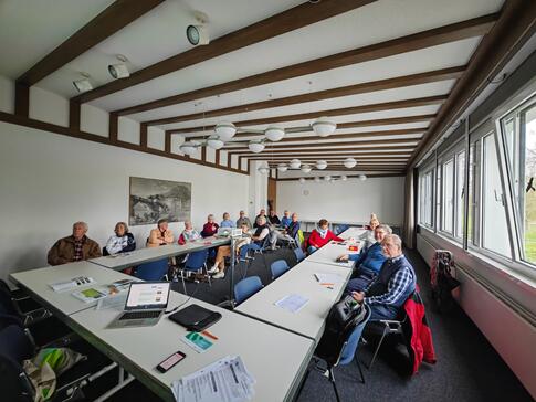 Die Teilnehmenden sitzen an einem großen, zusammengeschobenem Tisch im Raum und blicken in die Kamera