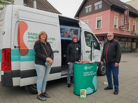 Johannes steht hinter dem Infostand vor dem Infomobil und neben ihm steht Frau Camus von der Bücherei, sowie ein Herr