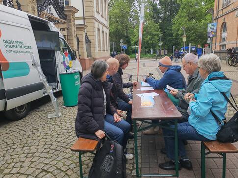 Besucher:innen sitzen auf Biertischen vor dem Infomobil und unterhalten sich mit unserem Referenten Johannes
