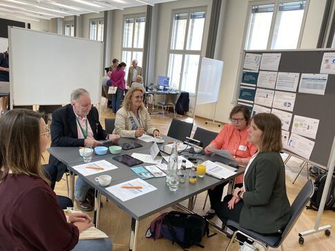 Theresa und die Besuchenden sitzen um einen viereckigen Tisch. Theresa erklärt Ihnen Digitale Gesundheitsanwendungen.