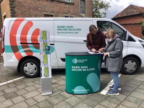 Petra steht mit einer Seniorin am Info-Stand vor dem Info-Mobil. Die beiden gucken auf ein Smartphone. Die Seniorin ist mit dem Finger an dem Smartphone.