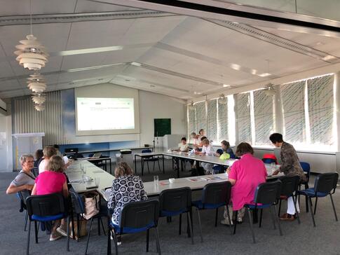 In einem großen Veranstaltungssaal im Neuen Kieler Rathaus sitzen in einem großen Tisch in U-Form rund 15 ältere Damen. Sie sind von hinten gezeigt. Im Hintergrund sieht man eine Projektionsfläche, wo allerdings nichts drauf zu erkennen ist.