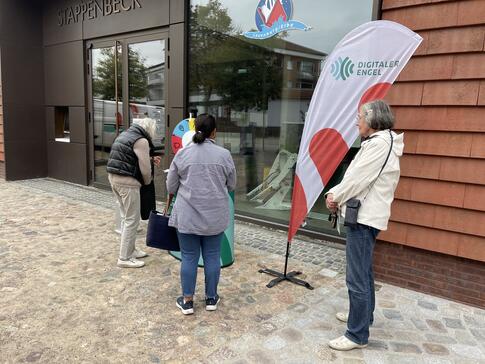 Drei Seniorinnen warten an unserem Infostand vor der örtlichen Bibliothek.