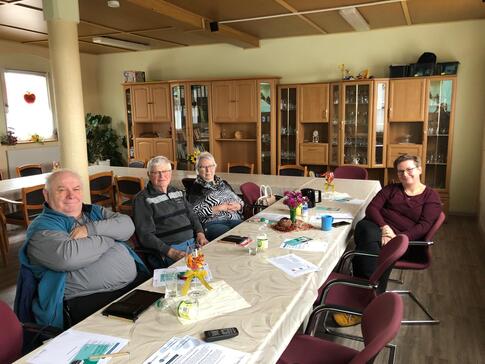 An einem längeren Tisch sitzen auf der linken Seite zwei Senioren und eine Seniorin, auf der rechten Seite sitzt eine Frau. Die vier Personen gucken nach vorne in die Kamera. Auf dem Tisch liegen verschiedenen Informationsmaterialien.