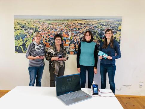 Petra (rechts) steht mit drei Frauen (links) an einer Wand vor einem Bild mit Ansicht von Wittichenau. Vor den Personen steht ein Tisch, auf dem ein Laptop und ein Smartphone liegen.