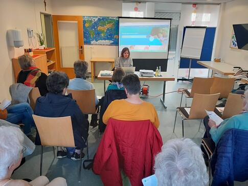 Mobilreferentin Theresa sitzt an einem Tisch und zeigt den Teilnehmenden Doctolib. Im Hintergrund ist die Doctolib-Internetseite auf einer Leinwand zu sehen. Die Teilnehmerinnen sitzen auf Stühlen und sind von hinten zu sehen. 