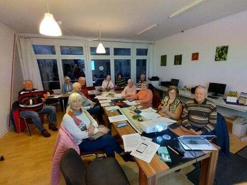 In einem Raum im MGH Buntes Haus sitzen vier ältere Männer und acht ältere Frauen an einem länglichen Tisch. Die meisten blicken nach vorne in die Kamera. Auf dem Tisch im vorderen Bildrand stehen ein Beamer und ein Laptop.