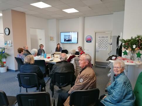 Im ROWI Stadtbüro sitzen insgesamt elf Personen. Im Hintergrund sitzen an einem Tisch Petra mit fünf älteren Menschen. Im Vordergrund sitzen auf Stühlen vier weitere Senior:innen. Die beiden Senior:innen ganz vorne drehen sich um und blicken in die Kamera.