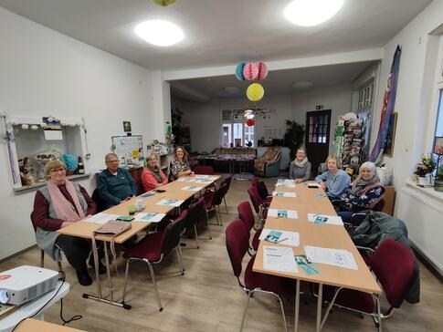 An zwei Tischen sitzen sieben Senior:innen und blicken nach vorne. Auf den Tischen liegen Materialien vom Digitalen Engel,