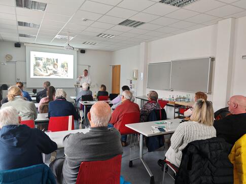 Mehrere Personen sitzen an Tischen im Raum und blicken nach Vorne zur Präsentation auf einer Leinwand