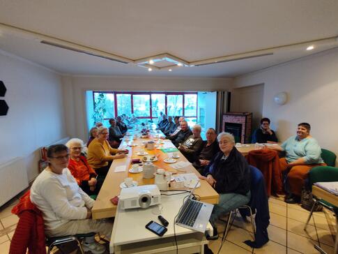 An einem langen, schmalen Tisch sitzen knapp 25 Senior:innen und blicken nach vorne. Am rechten Bildrand steht ein weiterer Tisch, an dem zwei Personen sitzen.