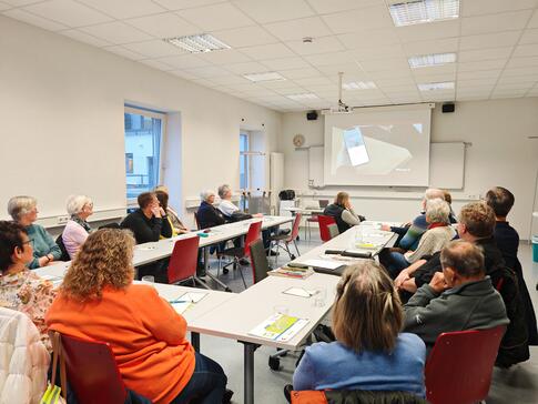 Mehrere Personen sitzen an einem zusammengeschobenen Tisch und blicken zur Präsentation auf einer Leinwand vorne