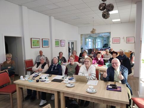 Im Plauderstübchen sitzen in vier Reihen knapp 20 Senior:innen und blicken nach vorne. Der Senior vorne rechts im Bild zeigt beide Daumen nach oben.