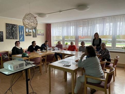 Die 8 Teilnehmenden sitzen an Tischen in einer U-Form. Theresa steht hinter ihnen und lächelt in die Kamera.