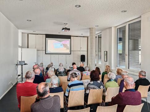 Mehrere Personen sitzen auf Stühlen im Raum und blicken nach Vorne auf eine Leinwand auf der ein Video abgespielt wird.
