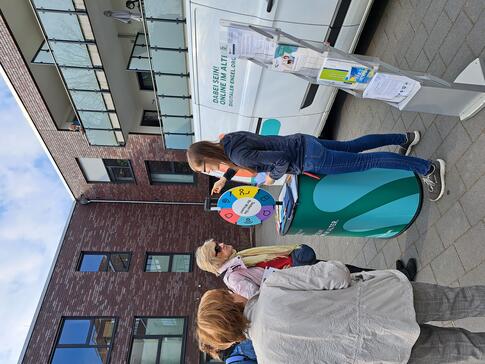 Petra steht mit dem Info-Stand vor dem Info-Mobil am Glücksrad. Drei Seniorinnen stehen um sie herum und eine Seniorin beantwortet eine Frage.