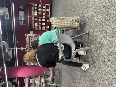Theresa und eine ältere Dame sind von hinten zu sehen und sitzen in einem Bibliotheksraum. Theresa zeigt ihr Dinge am Smartphone