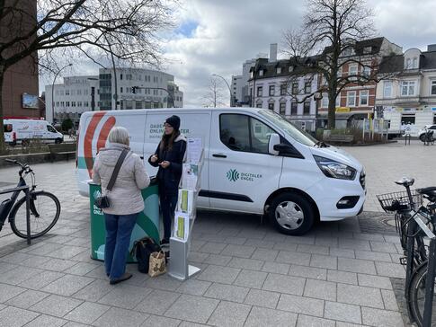 Petra steht mit einer Seniorin am Info-Stand vor dem Info-Mobil und ist mit ihr im Gespräch. Am linken und rechten Bildrand steht jeweils ein Fahrrad.