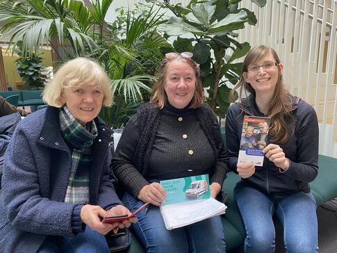 Petra (rechts) sitzt mit den beiden Freundinnen Astrid (Mitte) und Christine (links) im Atrium des KörberHauses. Petra und Astrid halten zwei Flyer in den Händen und Christine ihr Smartphone.
