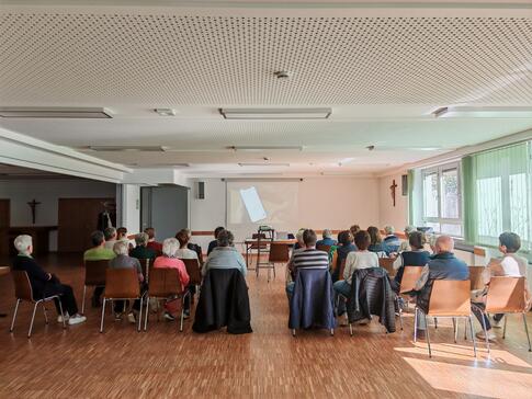Zahlreiche Personen sitzen auf Stühlen im Raum verteilt und blicken nach Vorne zu einer Leinwand.
