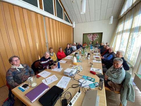 18 Senior:innen - davon die große Mehrheit Frauen - sitzen an einem langen Tisch. Sie gucken nach vorne in die Kamera. Auf dem Tisch liegen verschiedene Unterlagen.