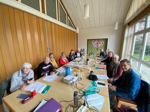 An einem großen, langen Tisch sitzen elf Senior:innen, davon ein Mann. Auf dem Tisch im vorderen Bildrand steht ein Beamer.