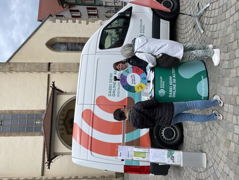 Theresa steht am Infostand vor dem Inbomobil und berät zwei Seniorinnen. Im Hintergrund ist ein Dom zu sehen.