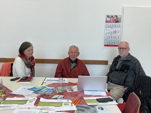 Die drei Teilnehmenden sitzen an einem mit Broschüren und Handouts gedeckten Tisch und lächeln in die Kamera.