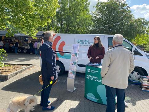 Petra (Mitte) steht an dem Info-Stand des Mobils und ist im Gespräch mit einer Frau (links, sie hält einen Hund an der Hundeleine) und einem Mann (rechts).