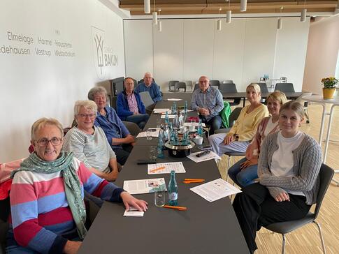 Neun Personen sitzen an einem langen Tisch und blicken in die Kamera. Auf dem Tischen liegen verschiedene Materialien.