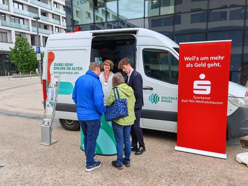 Johannes steht neben einem Berater der Sparkasse vor dem Infomobil und unterhält sich mit einem Ehepaar.