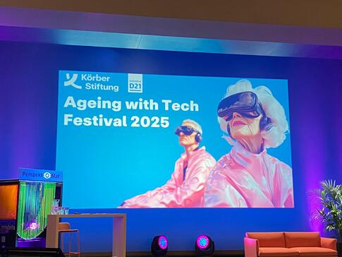 Eine Bühne im Theatersaal des KörberHauses. Eine Präsentation zeigt eine Folie mit der Überschrift "Ageing with Tech Festival 2025". Rechts ist eine KI-generiertes Bild mit einer älteren Dama und einer jüngeren Person, die eine VR-Brille tragen.