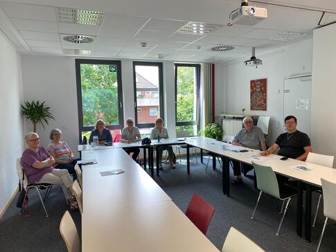 In einem Seminarraum sitzen an Tischen in U-Form fünf Frauen und zwei Männer. Alle blicken nach vorne in die Kamera. Die alle Teilnehmenden der Veranstaltung ließen sich fotografieren.