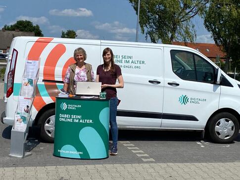 Petra (rechts) steht mit Seniorin Johanne (links) am Info-Stand vor dem Info-Mobil. Petra hält den Laptop von Johanne.
