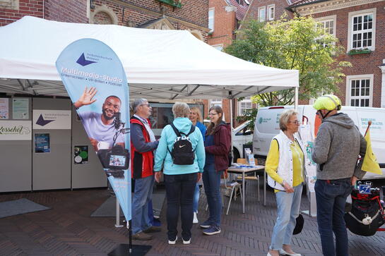Vor einem großen Pavillon steht Petra mit einem Ehepaar. Links davon steht eine Beachflag der Volkshochschule Meppen und rechts im Bild unterhält sich eine Frau mit einem Mann. Der Mann trägt einen Fahrradhelm.