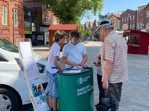 Zwei Schüler des Wahlpflichtkurses unterstützen einem älteren Ehepaar beim Info-Stand des Digitalen Engel.