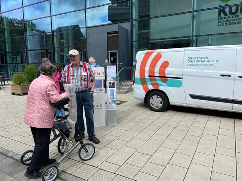 Eine ältere Frau mit Rollator steht mit einem Seniortrainer am Info-Stand neben dem Info-Mobil. Die beiden sind in einem Gespräch.