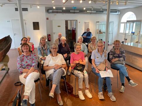 In einem Museumraum sitzen knapp fünfzehn Senior:innen in drei Stuhlreihen und schauen nach vorne in die Kamera.