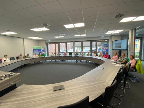 Im modernen Rathaussaal sitzen in einer großen ovalen Form mit Tischen ungefähr 17 Personen. Sie sind schwer zu erkennen, weil das Bild im Weitwinkelformat vor allem den Tisch vorne zeigt.