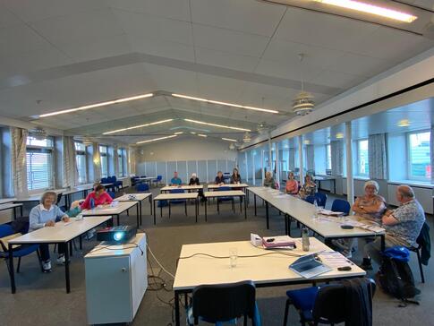 In einem sehr großen Raum sitzen ca. 13 Personen an Tischen, die in einer U-Form aufgestellt sind. Im Vordergrund steht ein Tisch mit einem Laptop und ein kleiner Schrank mit einem Beamer.