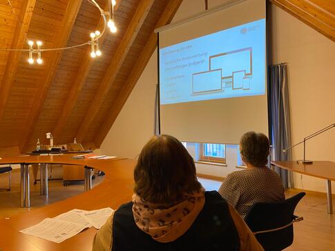 Zwei Damen sind von hinten fotografiert. Sie schauen nach vorne auf eine Leinwand, auf der verschiedene technische Geräte abgebildet sind.