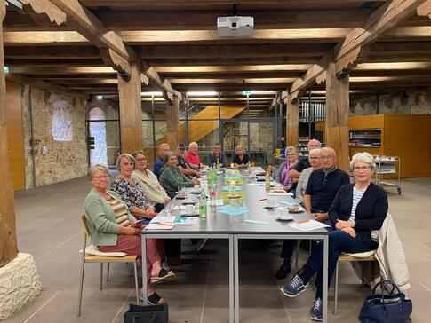 In einem Tagungsraum innerhalb der Burg Dagestein sitzen 16 Personen an einem langen Tisch. Sie blicken in die Kamera. Auf dem langen Tischen liegen Zettel und Broschüren.