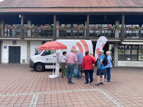 Das Infomobil inklusive des Standes steht vor einer Markthalle auf einem Marktplatz. Aufgrund des Wetters steht ein Sonnenschirm am Stand. Eine Gruppe von Senior:innen steht am Infostand.