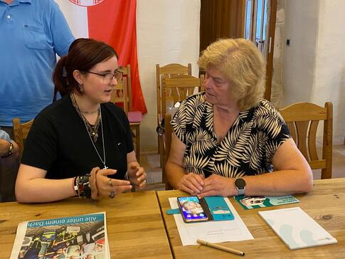 Schülerin Emma (links) unterhält sich mit einer Seniorin (rechts) an einem Tisch.